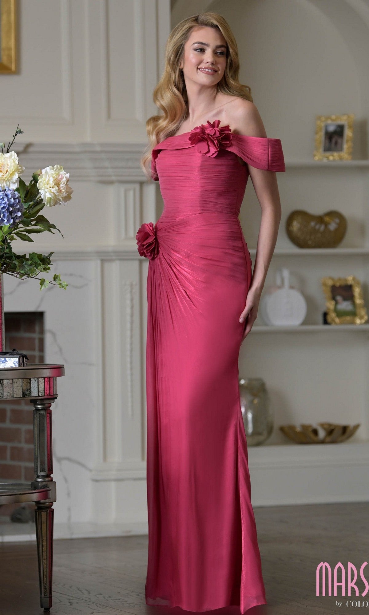 A woman poses indoors in the Marsoni M353 long formal dress in Oleander, featuring shimmer crinkled chiffon, an off-the-shoulder neckline, and 3D floral accents. She smiles near a fireplace with shelves in the background.